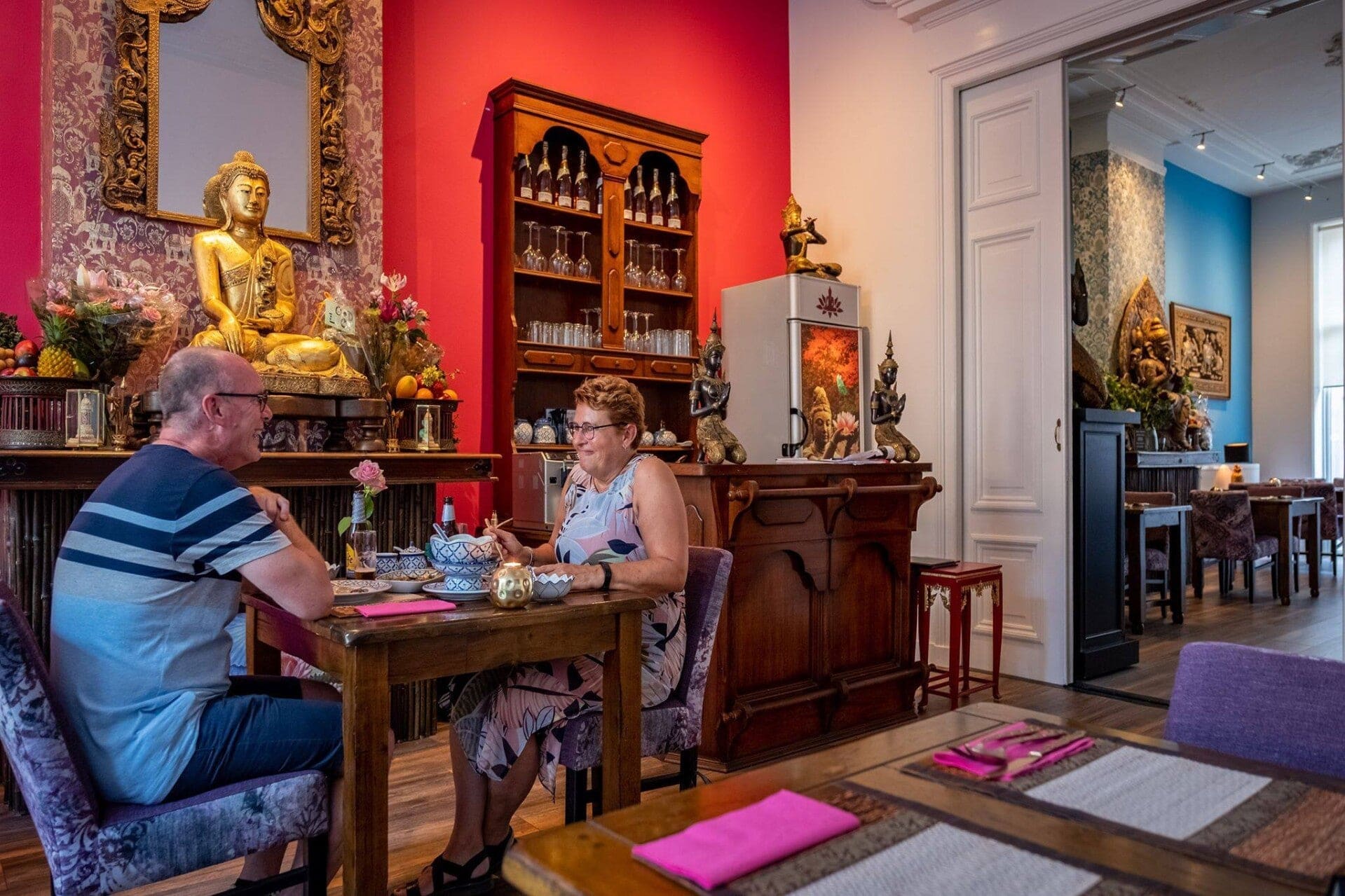 Gasten aan tafel in de roze zaal, met een gouden Boeddhabeeld tegen de rode wand.